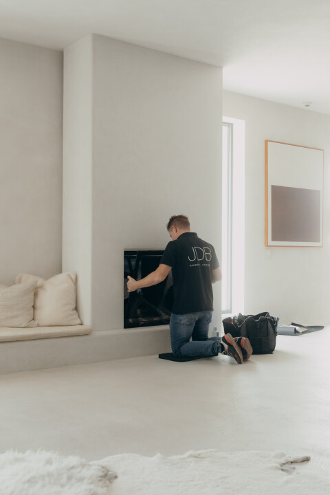 Een technicus van JDB Haarden voert onderhoud uit aan een ingebouwde haard in een minimalistisch ingerichte woonkamer. Professioneel haardonderhoud zorgt voor een veilige en efficiënte werking.