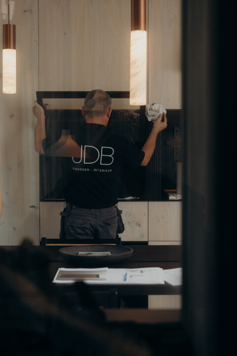Een technicus van JDB Haarden reinigt de glazen ruit van een ingebouwde haard in een modern interieur. Regelmatig onderhoud en reiniging zorgen voor optimale prestaties en een lange levensduur van de haard.